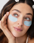 close up of womans face wearing light blue eye masks under her eyes. the masks say SOMETHING BLUE in white letters.