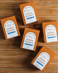 This image shows several brown-colored boxes with a white, dotted pattern and branding from "Old Whaling Co." Each box is labeled as "Shower Steamers," with the scent described as "Oatmeal Milk & Honey." The text on the packaging indicates that each box contains a "Set of 4" steamers with a net weight of 9.17 oz. The boxes are arranged on a textured surface resembling a woven or bamboo mat.