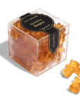 A transparent plastic cube containing amber-colored gummy bears labeled as "Off Hours Bourbon Bears" by Sugarfina. The black and gold label is affixed to the front, and a few gummy bears are placed outside the box on the surface, showcasing their translucent, golden appearance.