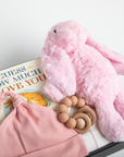 Pink plush toy, baby hat, and wooden teething ring on a book titled 'Guess How Much I Love You'.