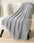Gray throw blanket with white fringe draped over a white armchair in a room.