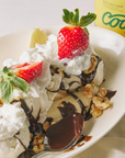 Ice cream sundae with whipped cream, strawberries, and chocolate sauce on a white plate, Coop logo in the background.