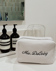 White toiletry bag with 'Mrs. DuCoing' branding on a bathroom counter with Aesop bottles.