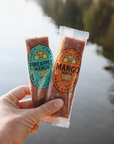 Two juice pouches held by a hand with a scenic background of water and trees.