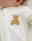 Close-up of a baby wearing a white zip-front bodysuit with a small embroidered teddy bear patch on the chest. The teddy bear is tan with a yellow bow, and the baby's ear and part of the face are visible.