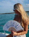 A blonde woman on a boat wearing white sunglasses and holding a white folding fan with 'bride to be' written on it, with turquoise water and a blue sky in the background.