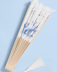 A traditional folding hand fan with white paper ribs and blue floral printed panels, featuring a white tassel at the bottom, set against a light blue background.