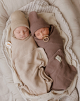 Two sleeping newborn babies swaddled in soft knit blankets and wearing knit hats, lying side by side in a bassinet. One swaddle is warm beige and the other is dusty mauve-brown, with matching pacifiers.