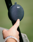 A close-up of a person's hand pressing a button on a round, dark blue Pund portable speaker mounted on a black pole against a soft-focus green background.