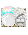 A boxed beauty gift set on a white background, containing a round iridescent eyeshadow palette labeled 'DIAMOND EYE,' a white scalloped makeup sponge, a white scalloped makeup brush, and a round silver compact with a label that appears to read Voluspa.