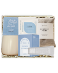 A curated wellness gift set in a white box with shredded paper filler, featuring a beige candle, blue and white packaging cards, a comfort pack pouch, and a gray cream packet on a white background.