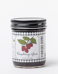 A glass jar of Finding Home Raspberry Jam with a black metal lid and a white label featuring a raspberry illustration, set against a plain white background.