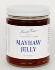 A glass jar of Trout River Goods Mayhaw Jelly, filled with dark amber jelly and featuring a white label with blue text and a white metal lid, set against a plain light background.