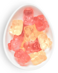Gummy bear candies in assorted translucent colors, including pink, red, orange, and pale yellow, arranged inside a white oval dish on a white background.