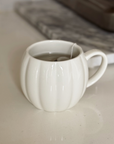 A white ceramic mug with a ribbed pumpkin-like design, filled with tea and a tea bag, sitting on a light countertop with a blurred kitchen background.