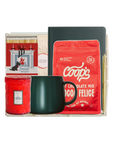 A curated gift basket set in a clear acrylic tray, featuring a red bag of Coop's Hot Chocolate Mix, a dark green ceramic mug, a red candle, a box of matches, and a black notebook, isolated on a white background.