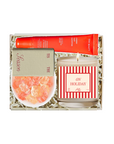 A curated gift set in a white box filled with shredded paper, featuring a red tube of hand cream, a striped candle labeled 'ON HOLIDAY', a small bowl of orange citrus peels, and a card reading 'Scented'.