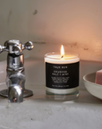A lit True Hue 'Crushed Salt + Mist' scented candle in a clear glass jar with a black label, placed on a bathroom sink next to a chrome faucet and a pink bar of soap on a white dish.