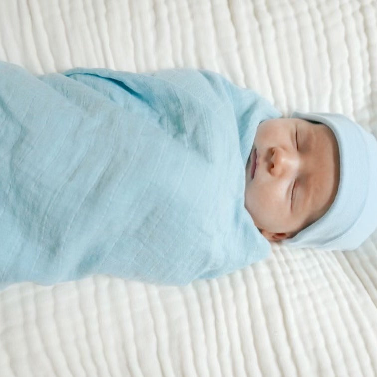 A baby wrapped in a blue snuggly cotton baby swaddle laying on a white muslin crib sheet.