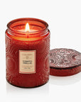 A red embossed glass jar candle with a lit flame, shown next to its matching patterned lid on a clean white background. The label reads Voluspa Tomato Trellis.