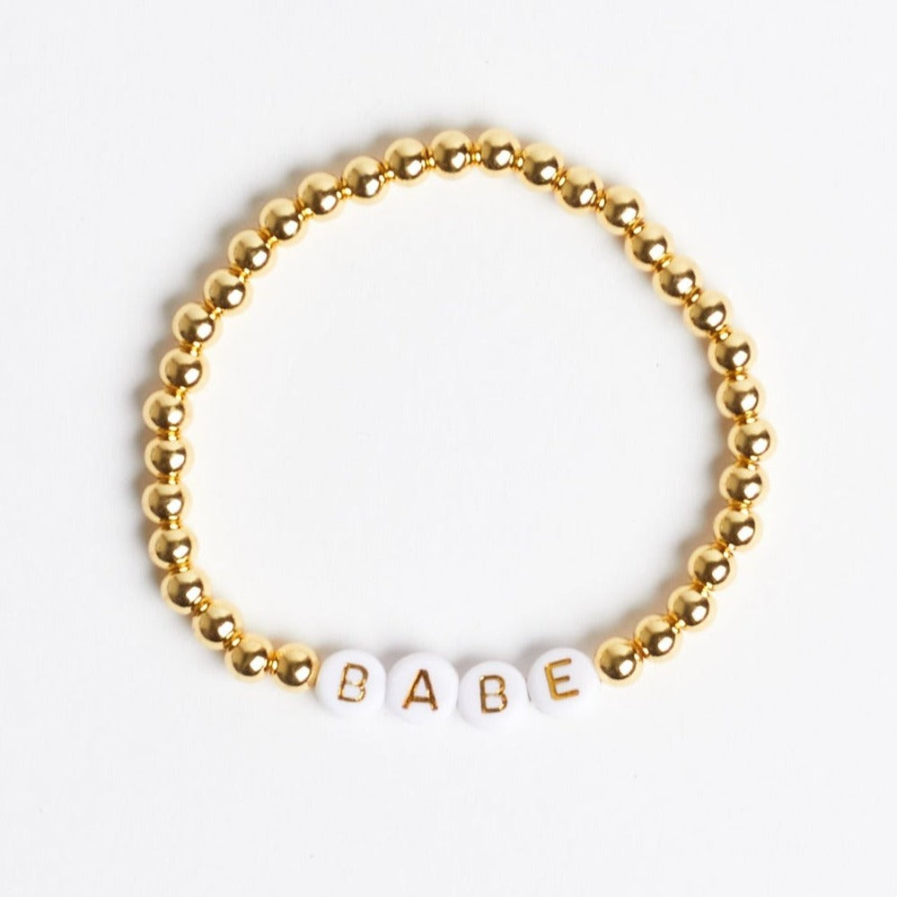 A gold beaded stretch bracelet with white beads that have gold text reading "B" "A" "B" "E" to spell out "BABE." Photographed against a white background.