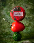 A stacked product display featuring a red round decorative container labeled 'VOLUSPA TOMATO TRELLIS' on top of a fresh red tomato, which is balanced on a green cucumber, set against a textured green background.