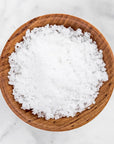 Coarse white sea salt crystals piled in a round wooden bowl on a light marble surface.