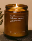 Brown candle labeled 'oakmoss + amber' with a flame on a dark surface.