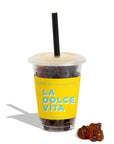 A clear plastic cup with a translucent lid and black straw, filled with dark gummy candies and wrapped in a bright yellow sleeve printed with the words 'LA DOLCE VITA'. A small cluster of brown gummy bears sits beside the cup on a white background.