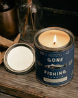 Candle in a 'Gone Fishing' can on a wooden surface with natural light.