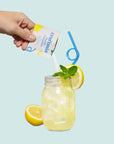 Hand pouring a drink mix into a glass with lemon slices and a straw on a light blue background