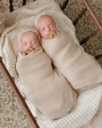 Two newborn babies swaddled in beige wraps with pacifiers, lying on a wooden crib.