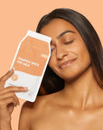 A woman with long dark hair holds a pouch of pumpkin spice oat milk next to her face against a solid peach background.