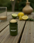 Green balm stick on a wooden surface with lemons and limes in the background