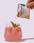 Hand pouring craftmix passionfruit powder into a pink drink with a mint leaf on a white background