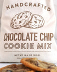 A cream-colored canvas bag printed with 'HANDCRAFTED CHOCOLATE CHIP COOKIE MIX' and an illustration of cookies, with chocolate chip cookies placed in front on a light marble surface.