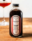 A bottle of Pappy & Company Bourbon Barrel-Aged Manhattan cocktail mixer sits on a wooden table with a blurred glass of the reddish-brown drink in the background.