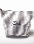 A gray and white vertically striped fabric pouch with a zipper closure and the name 'Tyra' embroidered in black script on the front, set against a plain white background.