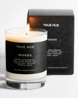 A lit True Hue Woods soy wax candle in a clear glass jar with a black label, standing in front of its black branded box on a clean white background.