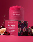 NCLA BEAUTY Hey Sugar exfoliating body scrub in a frosted jar, displayed on top of its pink packaging box with a fig and cherry pattern. The product is surrounded by fresh cherries and sliced figs on a solid pink background.