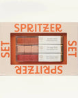 A boxed makeup spray set labeled SPRITZER SET, with a clear window showing three rectangular spray tubes in neutral and pink shades. The packaging is light beige with bold orange text, photographed on a clean white background.