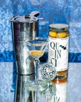 Cocktail shaker, glass with cocktail, and a bottle of Quin Sy on a reflective surface with a blue textured background.