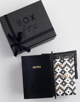 A black-themed stationery set including a notebook, a pen, a chocolate bar, and a coffee sachet, with 'BOXFOX' and 'THANK YOU' text on the packaging.