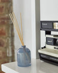 A blue glass reed diffuser bottle with natural reed sticks sits on a white shelf next to a vintage Polaroid camera. The background features a white wall and a blurred brick wall.