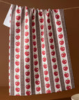 A kitchen curtain or hanging panel with a repeating red apple print on brown and white striped vertical bands, hanging from a rope against a warm brown background.