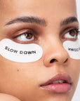 Close-up of a person's face with eye patches labeled 'SLOW DOWN' on a plain background