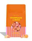 Sugarlina Champagne and Rosé gummy candy in a bright orange stand-up pouch with pink vertical stripes at the bottom and a clear window showing the gummies inside. A few gummies are placed in front of the bag on a white background.