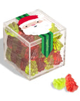 A clear acrylic cube container filled with colorful gummy bears, tied with a green ribbon featuring a Santa face. Two loose gummy bears sit in front on a white background.