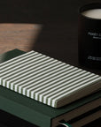 A striped notebook with white and dark green horizontal stripes sits on a dark green book, with a black candle labeled 'FORÉT CALME forest calm' in the background on a dark wooden surface.