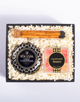A gift box set containing a Voluspa Crisp Champagne bottle cap, a pack of champagne buns, and a square box of 'Champagne bears' gummy bears, all arranged in a black tray filled with shredded white paper on a white background.